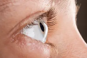 A close-up view of a person's eye with a contact lens and eyelashes, possibly a woman, has a white color on the cornea and a black color on the pupil, with a small tear drop in the corner of her eye.