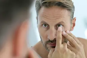A man with brown eyes is looking at his reflection in the mirror and trying to put a contact lens in his eye while holding his eye with his right hand.