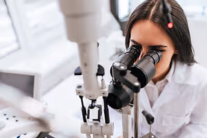 A woman is looking through an ophthalmoscope, possibly in a laboratory.