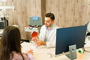 A doctor in a white coat explains an eye model to a woman in a medical office.