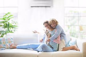 A family of two women and a child with a dog sitting on a white couch in a living room with an air conditioner, plant, and lamp.