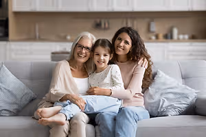 A family of three, an elderly woman, a young woman, and a young girl, sitting on a couch and smiling while looking at the camera.