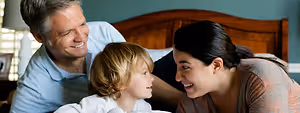 A family of three is smiling and looking at each other while lying on a bed in a bedroom