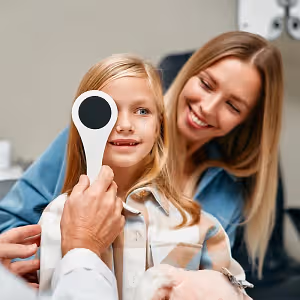 Girl getting her eye checked by an optometrist with her mother smiling in the background