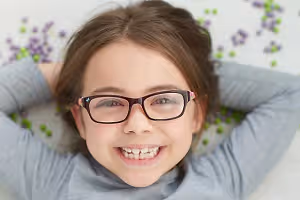 A smiling young girl with glasses lying on a white surface with colorful beads behind her