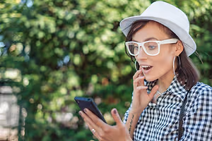 A woman wearing a white hat, glasses, and a checkered shirt is holding a cell phone in her left hand and seems to be talking to someone on the phone. She is wearing a ring on her left hand and an earring on her right ear.