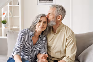 A man and woman are sitting on a couch, the man is kissing the woman's forehead