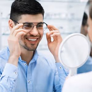 A man is adjusting his glasses and smiling while looking at a mirror