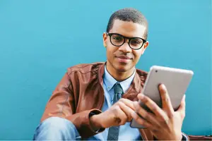 Man sitting against a blue wall while holding a tablet computer and smiling
