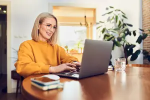 A woman with blonde hair is smiling and using a laptop on a wooden table.