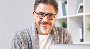 A man with a white beard wearing glasses and a gray sweater is smiling at the camera