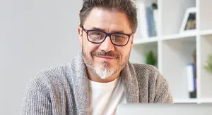 A man with a white beard wearing glasses and a gray sweater is smiling at the camera