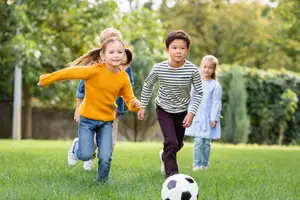 A group of kids are playing soccer on a grass field, one of them is a girl in a yellow sweater kicking a ball