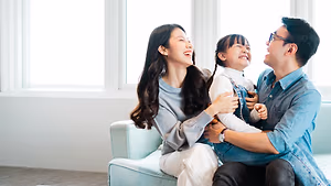 A family of three, a woman, a man, and a young girl, sitting together on a couch and laughing at something in front of them.