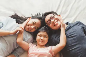 A smiling family of three lying on a bed with their hands on their heads