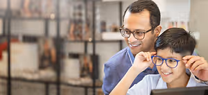A man and a boy are standing in front of a monitor, smiling, with the boy wearing glasses and the man holding the boy's glasses.