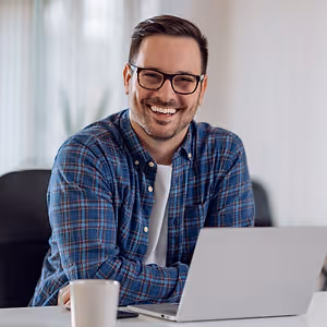 A man with glasses sitting on a chair in front of a laptop, smiling