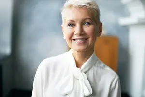 Portrait of a woman with short blonde hair, wearing a white blouse with a bow, smiling and standing indoors.
