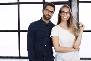 A smiling couple wearing glasses posing for a photo in front of a glass window.