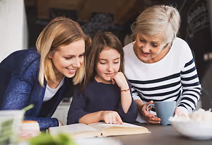 A family sitting at a kitchen table, with a young girl reading a book and the adults smiling and looking at her