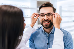 A man wearing glasses is having his eyeglass frame adjusted by a woman in a lab coat.