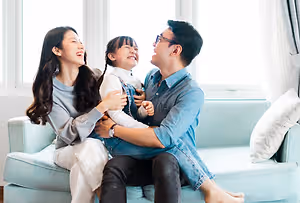 A family of three sitting on a blue couch with a young girl laughing and looking at the camera