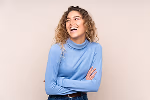 A woman with curly hair, smiling, and posing with arms crossed in a blue turtleneck and jeans against a beige wall
