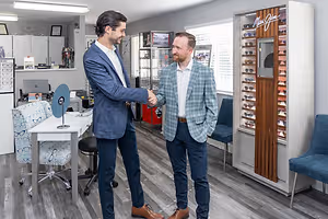 Two men in formal wear are shaking hands inside an office. One man is wearing a blue blazer and the other is wearing a checkered blazer. Behind them is a desk with a laptop and a cabinet with some items on top. There is also a glass display case with sunglasses on the right side of the room.
