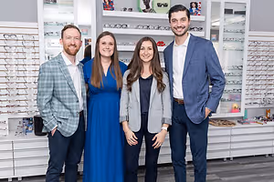 Four people in suits posing for a photo in an optical shop with cabinets filled with glasses behind them