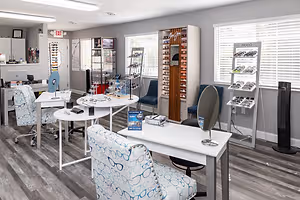 An interior shot of an optical shop with several tables and chairs, one with an eyeglass display, and a shelf with glasses on display. The shop is well-lit with ceiling lights and natural light coming from the windows with blinds.