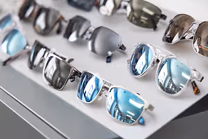 Various pairs of sunglasses with different designs and colors are displayed on a white surface, possibly in a shop or store, with reflections of buildings and lights on the lenses.