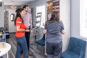 Three people are inside a shop, one of them is wearing a red shirt, and they are standing near a display of sunglasses and chairs