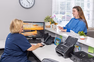A woman sitting at a desk talking to another woman in a blue and white striped shirt