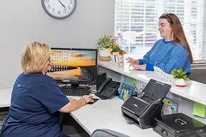 A woman sitting at a desk talking to another woman in a blue and white striped shirt