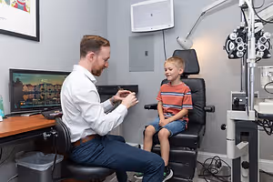 An adult male doctor sitting on a chair beside a boy on a chair with a monitor on the desk beside him.