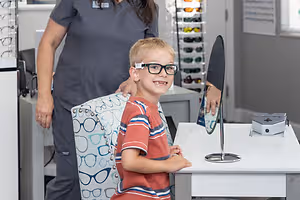 A woman wearing gray scrubs stands behind a young boy in a red and white striped shirt who is wearing glasses and sitting in front of a white desk with a mirror and a camera.