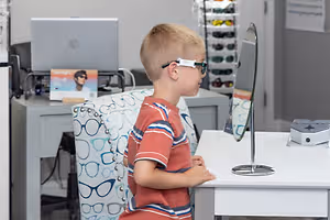 A boy in a red striped shirt is sitting at a table looking at himself in a mirror while wearing glasses.