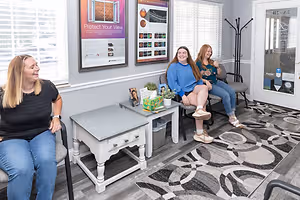 Three women sitting in a waiting room while one woman looks at the camera and smiles.