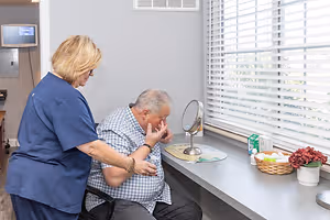 A nurse in a blue uniform is examining a patient sitting in a chair near a window with white blinds and a basket of flowers on a counter.