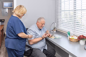 A man is sitting in a chair in a medical room while a woman stands next to him, holding a magnifying glass and smiling. The room is well-lit with a white wall, a window with blinds, and a desk with various items including a mirror, a basket, and a flower vase. The man is wearing a bracelet and a checkered polo shirt.