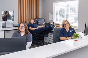 Three women in a reception area with one woman sitting in a chair with her legs up on the desk, another woman sitting at a desk, and the third woman sitting at a reception desk with a potted plant on it.