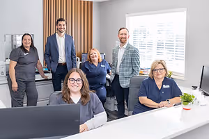 A group of people are posing for a photo in an office room, smiling and standing near a desk and a window. There are several items on the desk, such as a monitor, a potted plant, and a white desk.