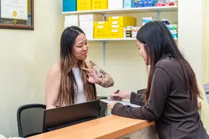 A woman with tattoos is speaking with another woman in a clinic. A woman in a brown jacket is holding a pen and clipboard and seems to be writing on it. A shelf with several boxes is behind them. A laptop is on the desk in front of them.