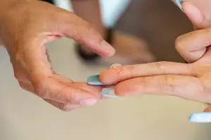 Two hands are holding a small contact lens between them. The lens is clear and transparent, and it appears to be placed on the tip of the index finger. The other hand is holding the lens with its thumb and index finger, and the other fingers are supporting it. The image is blurry, and it seems like the lens is being examined closely.