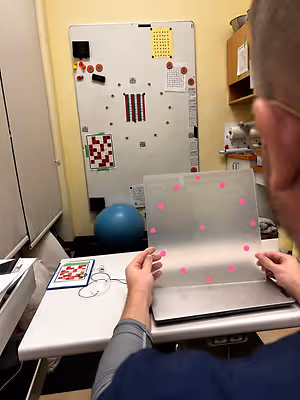 A man wearing glasses and a blue shirt is holding a clear plastic sheet with pink dots in front of a whiteboard in an indoor setting.
