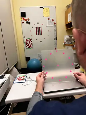 A man wearing glasses and a blue shirt is holding a clear plastic sheet with pink dots in front of a whiteboard in an indoor setting.
