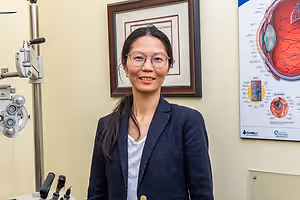 A smiling woman wearing glasses stands in front of an eye examination setup in a clinic