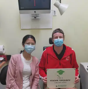 A man and woman wearing face masks sit in a medical office holding a certificate that says I'm a Vision Therapy Graduate