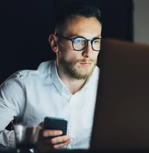 A man in a white shirt and glasses holds a smartphone while looking at a laptop screen in a dimly lit room