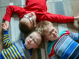 Three young boys wearing glasses, lying on a wooden floor with smiles on their faces, one with his hands clasped together, another with a hand resting on his chest, and the third with his hand on his forehead, possibly posing for a photo in a room.